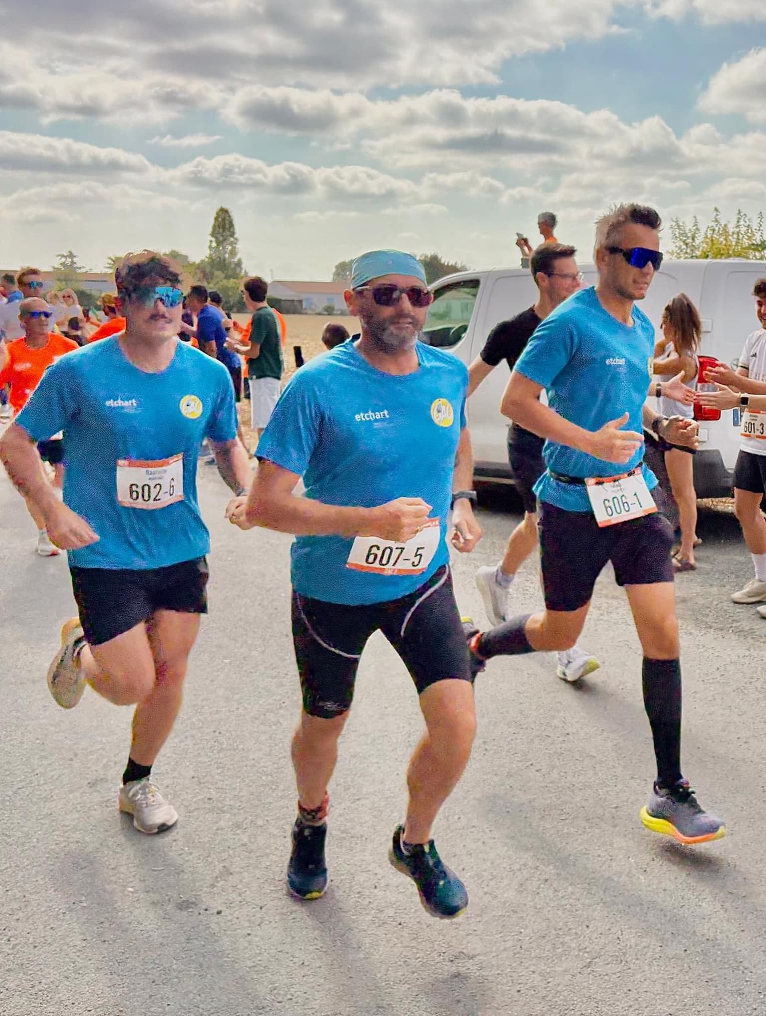 Trois coureurs du CAC en tee-shirt bleu et dossard courant en relais sur une route, encouragés par le public